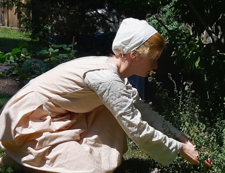 A white woman with blonde hair covered by a small white cap crouches beside a herb garden. She wears a  long Pink sleeveless  dress over a long-sleeved cream shirt